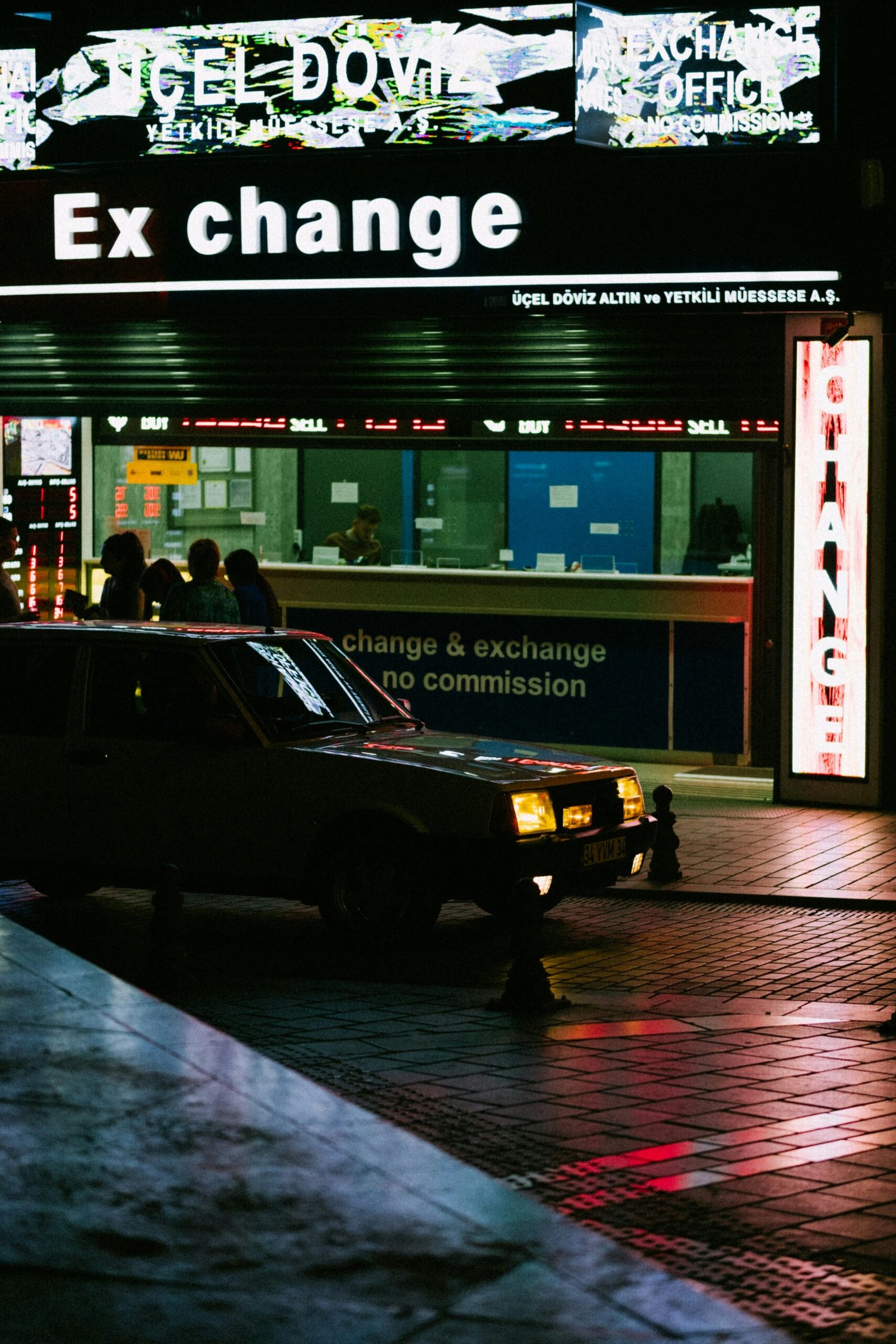 Illuminated currency exchange storefront at night with street reflections and a parked vehicle.