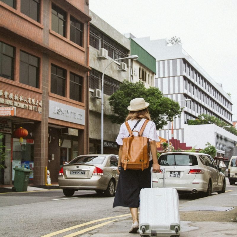 pexels-photo-119777-119777 A woman with a suitcase and backpack walks down a busy city street on a sunny day.